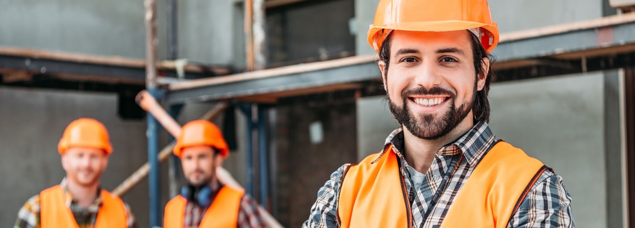 knappe bouwondernemer staat met zijn armen over elkaar op een bouwplaats met zijn collega's op de achtergrond